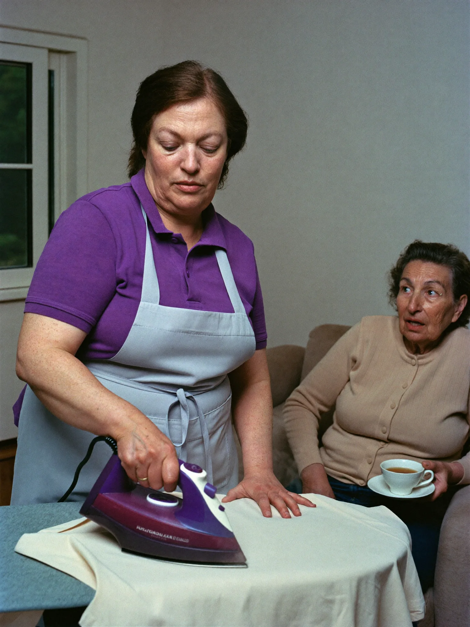 Haushaltshilfe bügelt, die Seniorin sitzt mit einer Tasse daneben, Einsatz in Simmozheim
