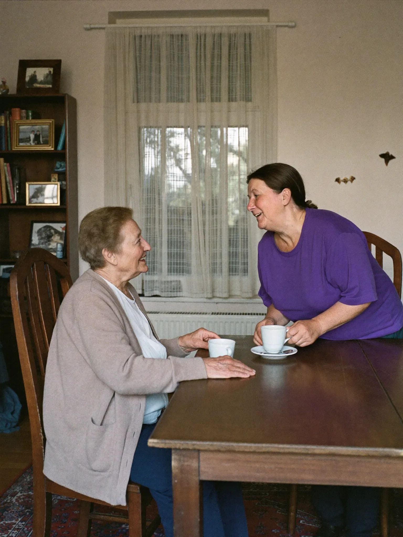 Haushaltshilfe und Seniorin am Esstisch, dahinter ein Bücherregal in Schömberg